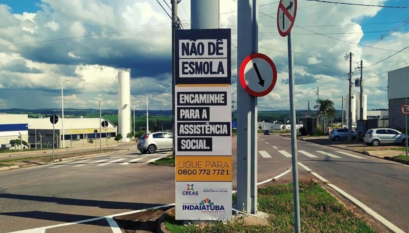 Placa de sinalização vertical em um cruzamento de Indaiatuba com os dizeres "Não dê esmola, encaminhe para a assistência social", exibindo o telefone 0800 do CREAS e a logomarca da Prefeitura, sob um céu nublado.